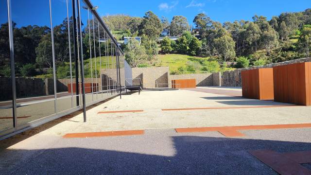 A glass-fronted building borders a courtyard with low rust-coloured forms, paving lines, stone walls and a green hillside beyond.