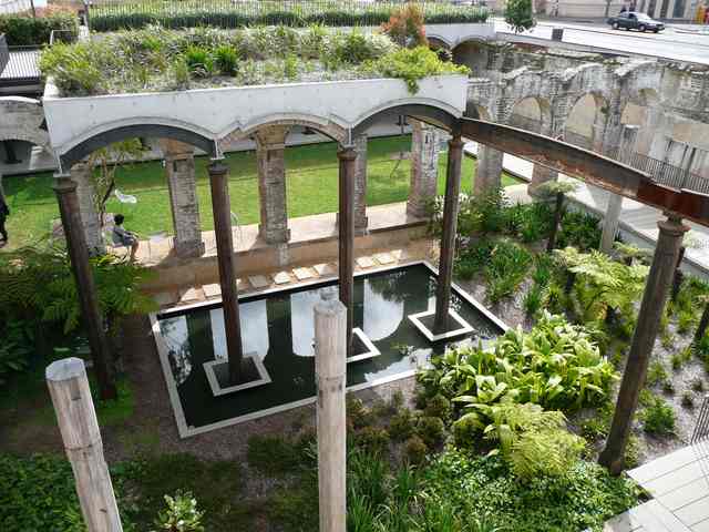 Looking down into a planted sunken garden, with a rectangular pond beneath brick arches and metal beams; the street is visible above.