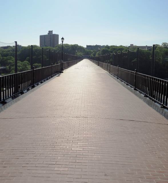 A long brick walkway recedes between railings, mesh fencing and lamps toward trees and Bronx buildings.