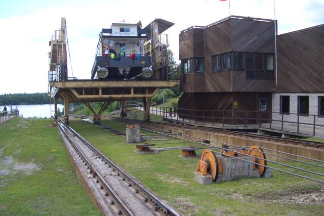 A houseboat with several distant passengers rests on a raised steel railway carriage beside a timber-clad building, with rails, cables and grass underneath.