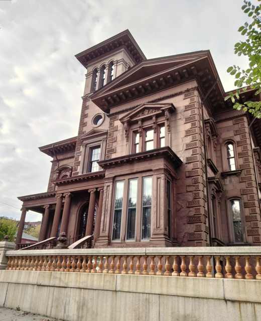 Looking up at the brownstone façade, carved window surrounds, columned entrance and tower above a stone balustrade.
