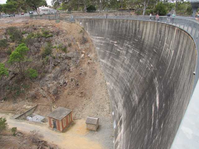 The tall concave concrete wall curves above small utility buildings, with a railing and visitors along its crest.