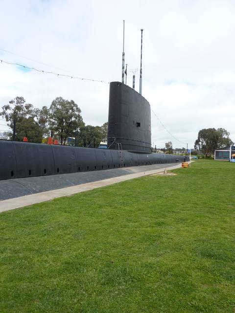 A tall black fin and slender masts rise from the long casing beside a paved path and lawn.