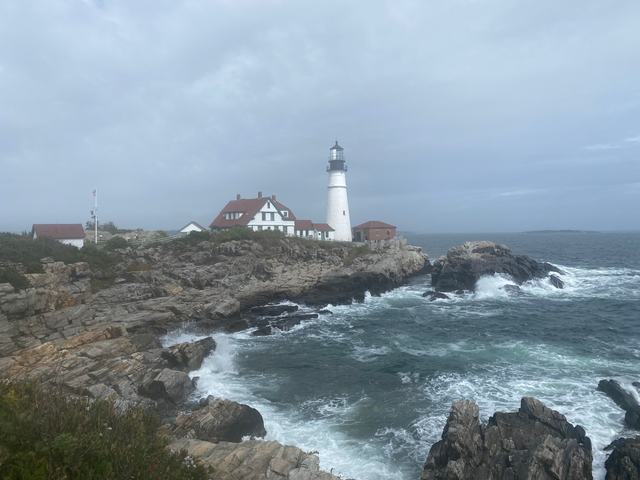 The keeper house and white tower stand on a rocky headland, with waves below and mist behind them.