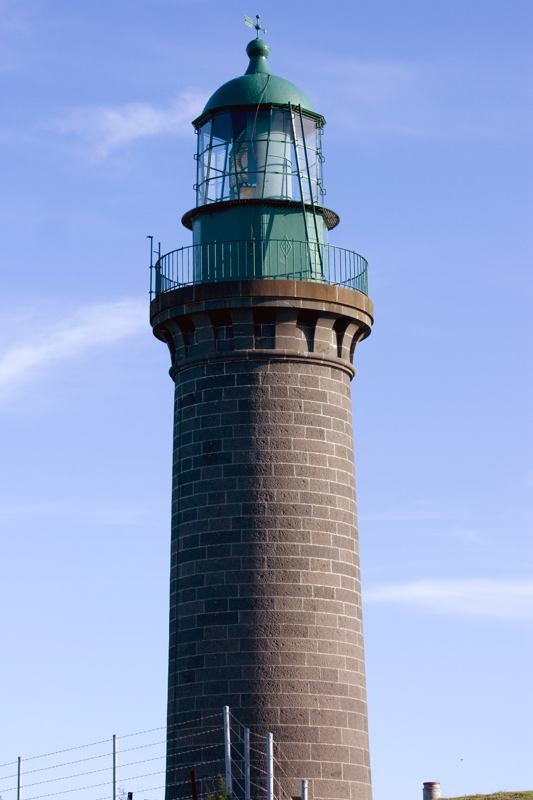 A dark masonry lighthouse with green lantern and railing rises behind a fence against a blue sky.