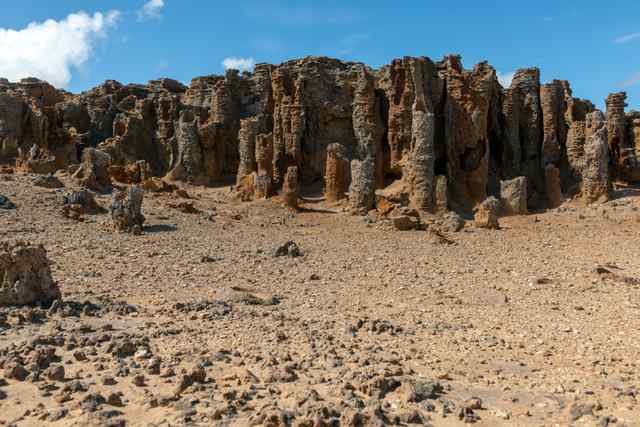 Tall corrugated tan and brown columns stand above loose stone, with low shrubs to the left and a blue, clouded sky.