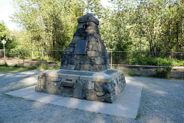 A stone commemorative cairn with attached plaques stands in public grounds beside a fenced railway.