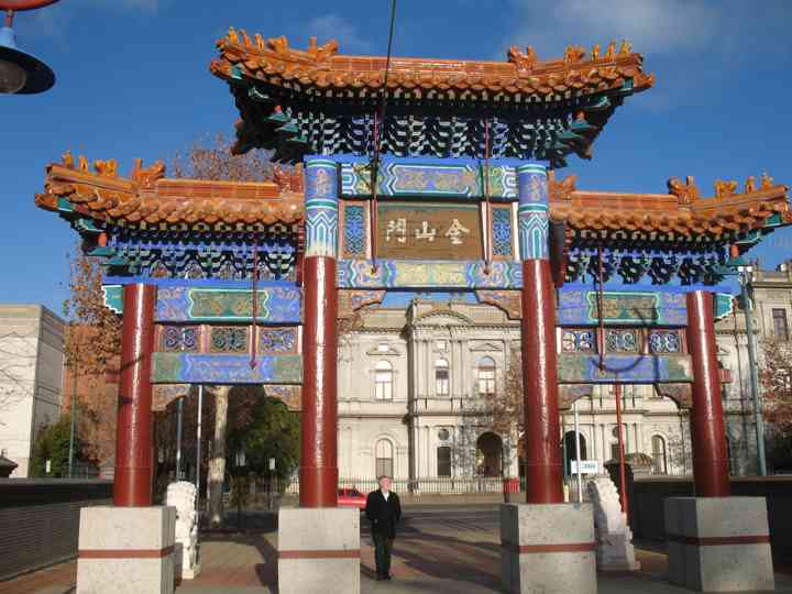 An ornate gateway with orange tiled roofs, red columns and blue-green decoration frames the street and buildings beyond.