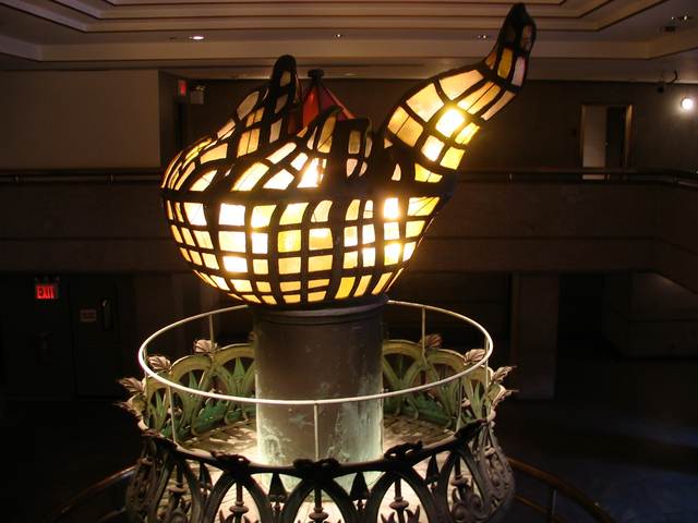 An irregular flame of glowing amber and pale glass panels sits above a metal handle and circular railing inside a gallery.