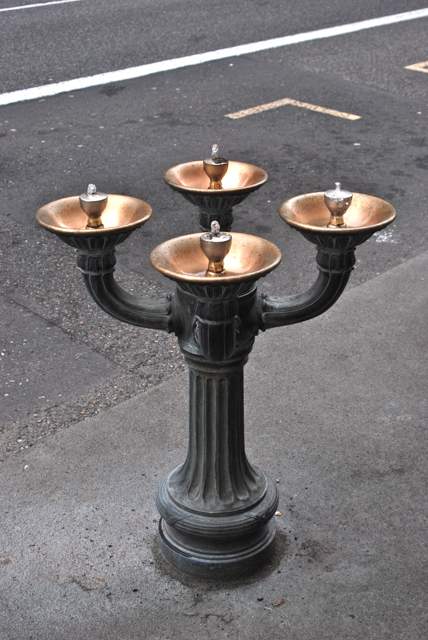 Four bright bowl interiors surround a dark ornamental stem, with small water jets above the nozzles.