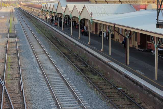 Several railway tracks run beside the long covered platform, where small groups of passengers wait.