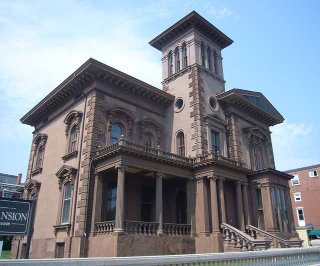 A brownstone mansion with a tall central tower, arched windows, columned porch and stone entrance steps.