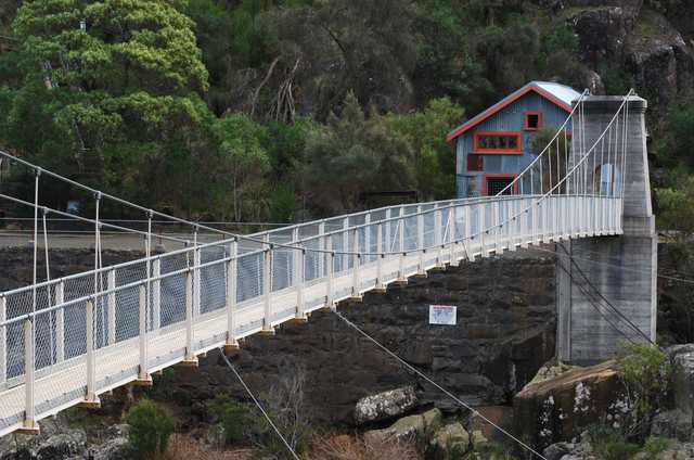 A pale suspension footbridge crosses a rocky gorge toward a concrete tower and small blue-and-red building against a steep wooded bank.