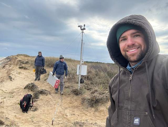 Three people with field equipment stand beside a camera pole on a sandy bluff.