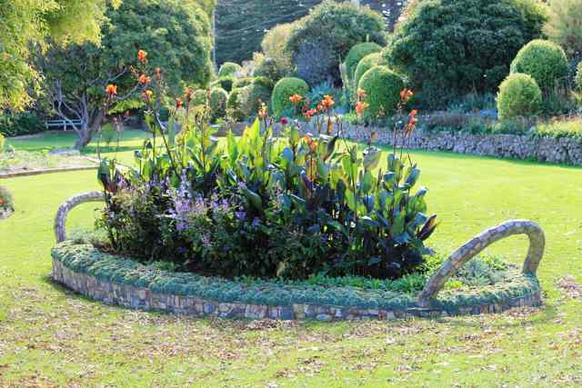 Orange and purple flowers rise from a low stone-faced garden bed with two curved handles, surrounded by lawn and clipped shrubs.