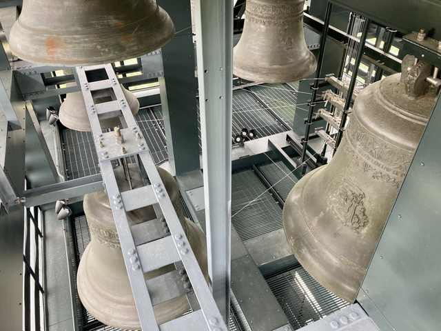 Large bronze bells hang among steel beams, grated platforms, wires and levers inside a tower.
