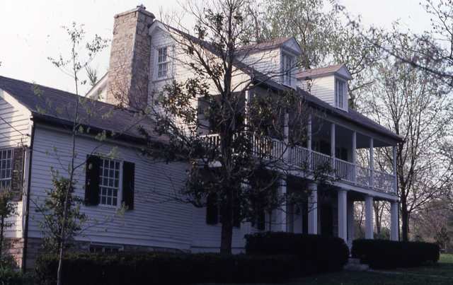 White siding and a two-story porch are partly screened by trees beside a stone chimney.