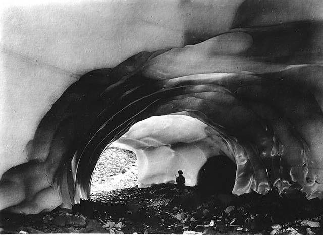 A silhouetted hiker stands beneath a layered ice arch, with daylight beyond.