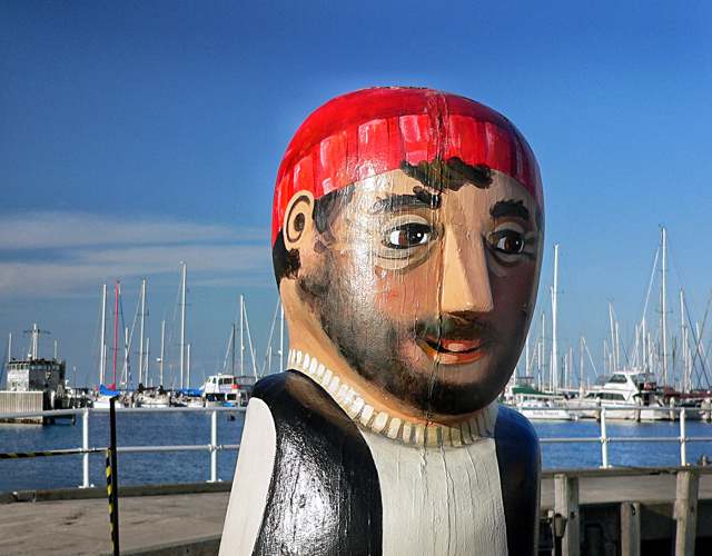 Close view of a bearded painted wooden figure with a red cap; vertical lines in the timber cross its face, with boats behind.