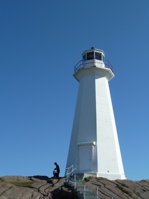 A tall white concrete tower and glazed lantern stand on rock against a blue sky; two small visitors are near its base.