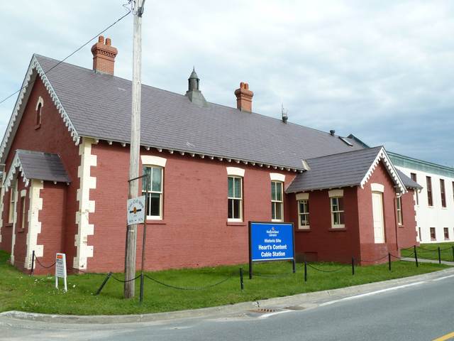 A red-brick building with white trim and a small gabled entrance beside a road; a white extension stands behind it.