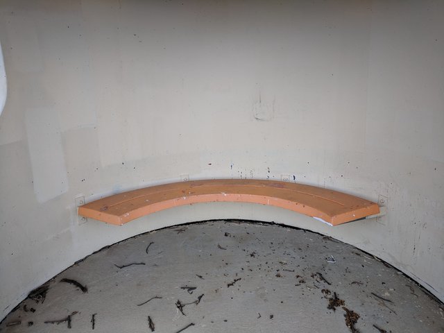 Curved orange bench attached to a concrete shelter wall, with a worn surface and the floor below.