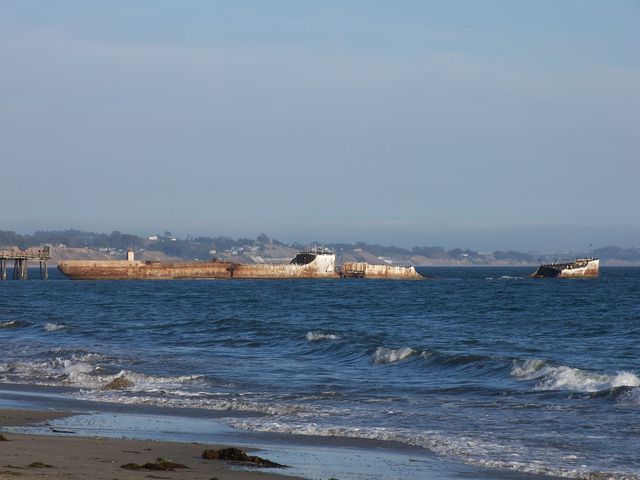 The broken concrete hull extends across a blue-water view with shoreline behind and a short section of pier at left.