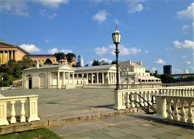 Pale colonnaded waterworks buildings stand beyond a paved terrace and railings, with an art museum uphill and a streetlamp in the foreground.