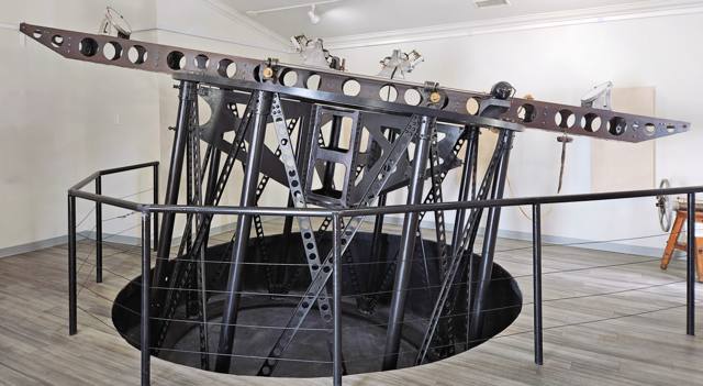 A long perforated beam carrying mirrors above a tall black metal support inside an exhibition room.