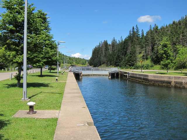 A water channel runs beside a paved lockside toward gates and railings, with trees on either side.