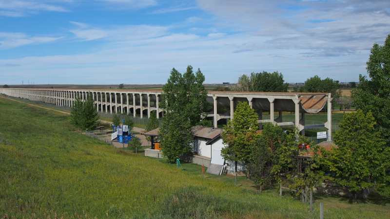 An exposed curved concrete channel end stands above supporting columns, with trees, small buildings and a playground in the foreground.
