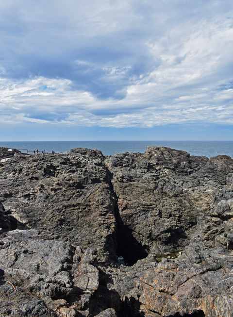 Rough coastal rock surrounds a deep opening, with ocean and clouds beyond.