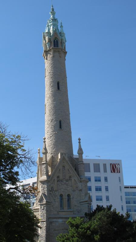 A tall pale-stone tower rises above a gabled, buttressed base, with an ornate crown, trees and a modern building behind it.