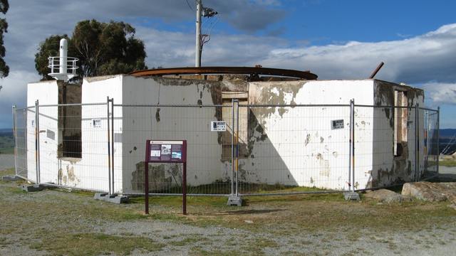 White, weathered walls stand without a roof behind temporary fencing; a rust-coloured metal ring is visible above them.
