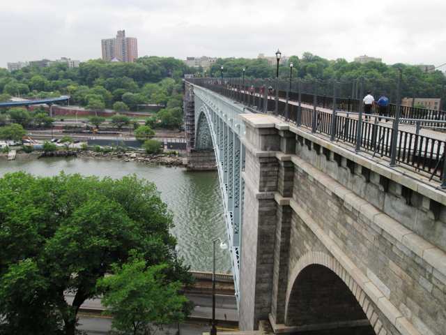 A steel arch meets a stone pier above the Harlem River, with railings, lamps and distant people along the deck.