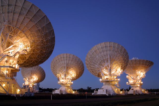 Illuminated radio dishes beneath a deep blue sky, with one large dish partly outside the original frame.