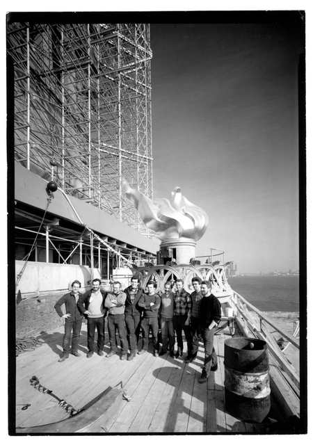Workmen stand beside a torch with a solid sculpted flame, with scaffolding behind them and New York Harbor at right.