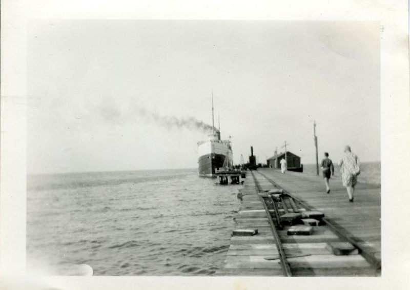 Black-and-white photograph of people walking along a wooden dock toward a steamer, with rails running beside the walkway.