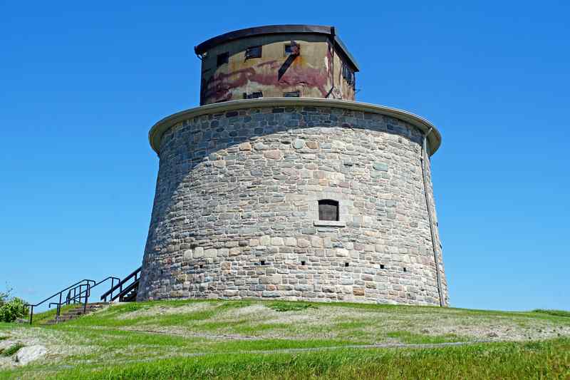 A round stone tower on a grassy rise, topped by a smaller angular concrete structure with observation openings.