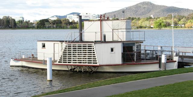 A white paddle steamer moored at a jetty, with its complete hull, paddle housing, upper cabin and funnel visible.