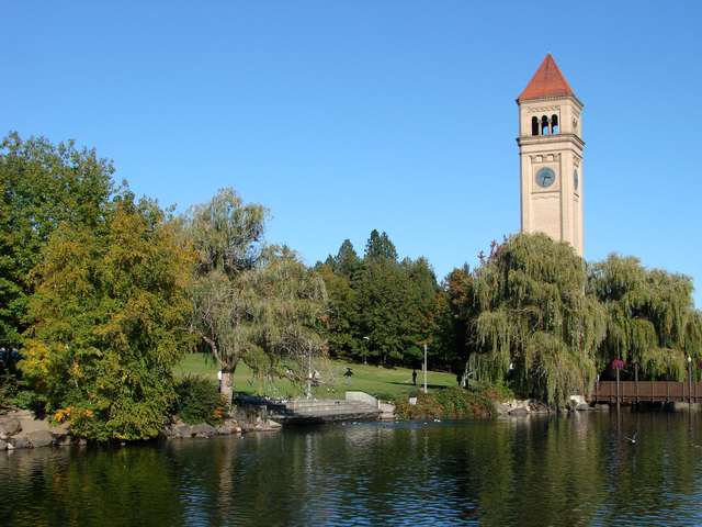 A red-roofed clock tower rises above green trees and drooping willows across a river, with a lawn and small distant figures below.