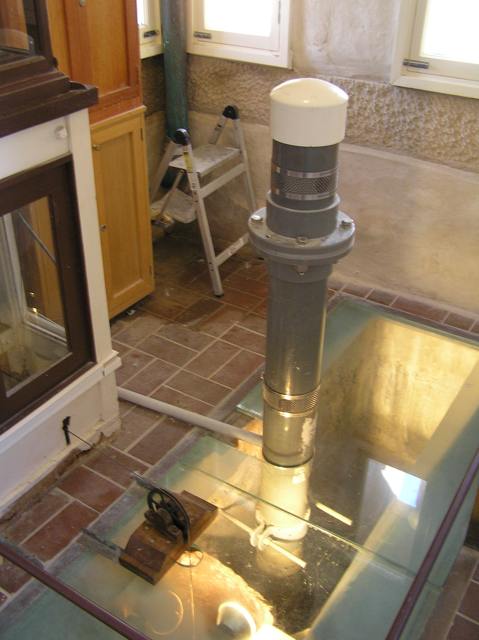 A white-capped grey cylindrical instrument rises through a glass floor panel in a stone-walled room.
