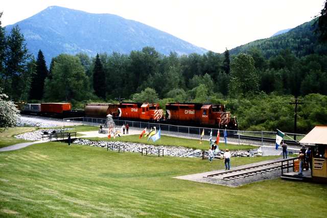 A freight train passes the memorial grounds, where a small stone cairn, flags and visitors stand below wooded mountains.