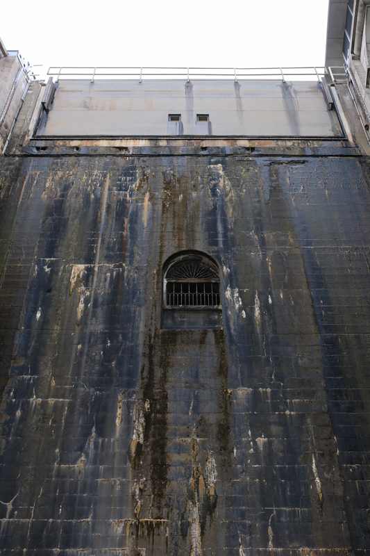 A dark concrete wall with pale vertical marks and a barred arched opening rises to a broad metal gate and railing.