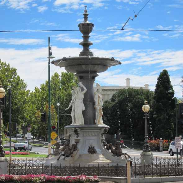 Pale sculpted figures stand beneath the fountain’s dark upper bowl, with water jets, a low iron fence, flowers and surrounding road traffic.
