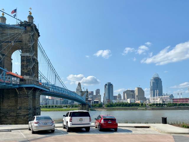 The suspension bridge crosses the river toward a city skyline, with parked cars in the foreground.