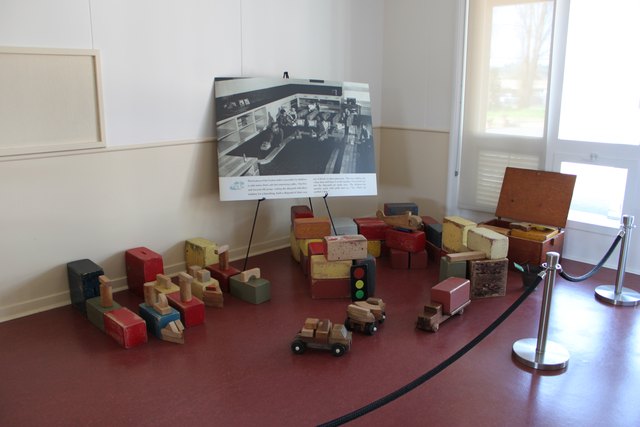 Wooden blocks and toy vehicles sit behind a rope barrier, with an interpretation panel on an easel beside them.