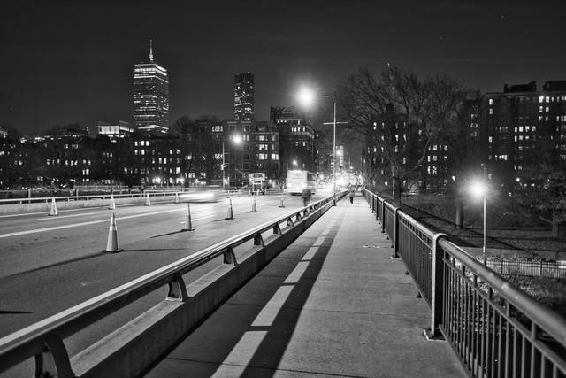 Black-and-white view down a bridge sidewalk toward illuminated buildings; a rail separates pedestrians from blurred traffic.