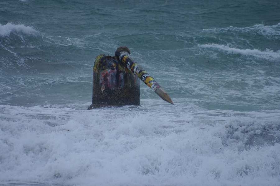 A painted concrete mast lies diagonally across its cylindrical base amid rough water.