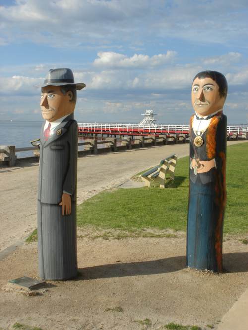 Two tall painted wooden figures stand near the waterfront: one in a grey suit and hat, the other in a ceremonial robe and chain.